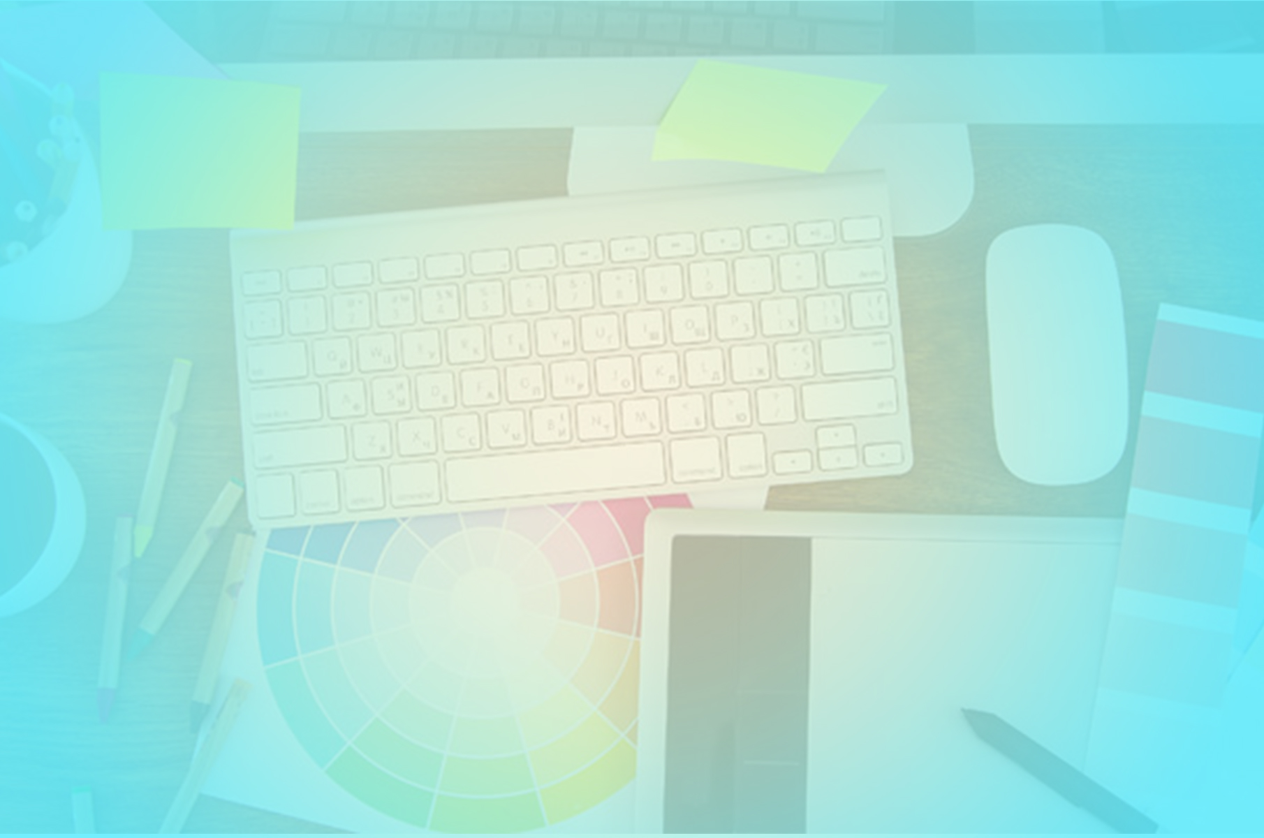 Overhead view of a designer desk with keyboard, mouse, colour wheel, sticky notes and drawing tablet, tinted in cool blue-green studio light
