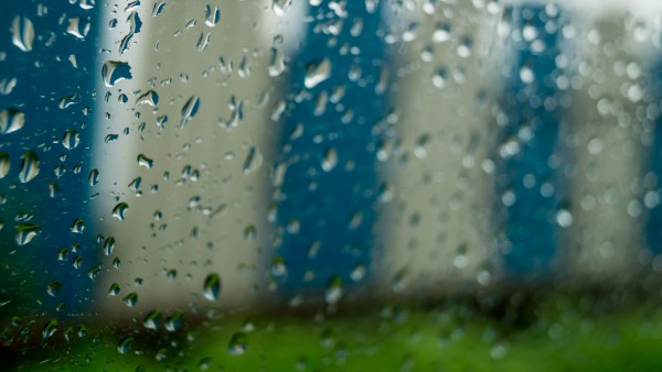 Close-up of raindrops on a window with blurred blue and white stripes and a band of green below, cool cinematic tones